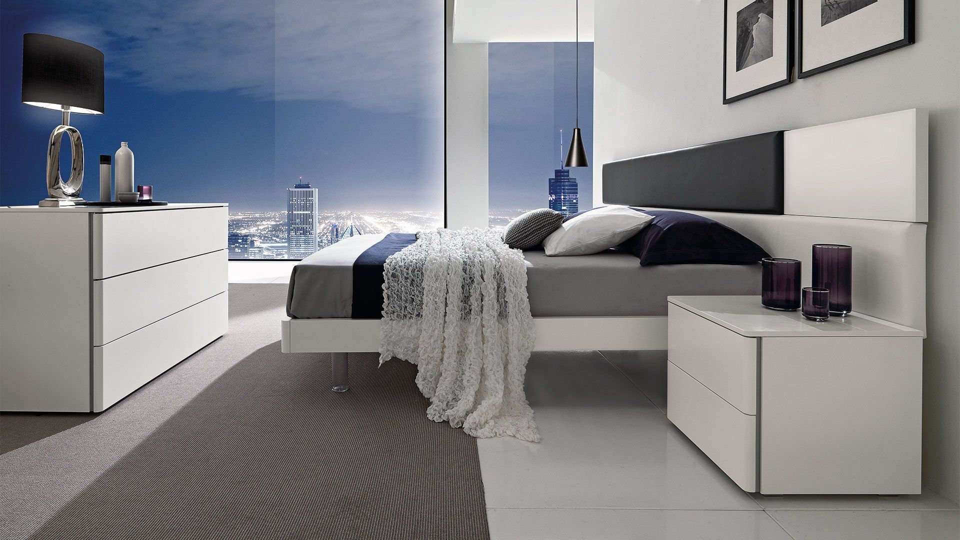 Camera da letto moderna con mobili bianchi, vista sulla città e cuscini decorativi.