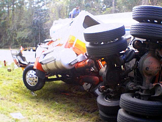Rolled-over semi-truck on its side; orange cab, white tanker, and multiple tires visible on grass beside road.