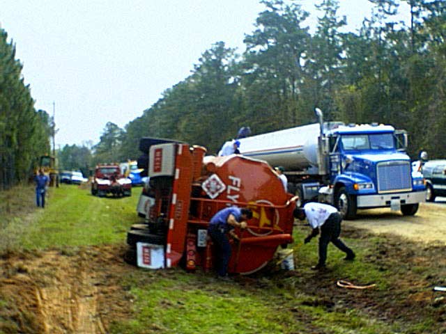 Overturned fuel tanker truck in ditch, emergency responders assess scene on roadside. Blue truck, red tanker.