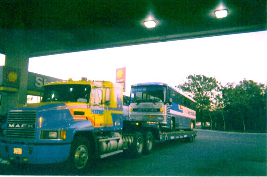 Blue and yellow Mack tow truck towing a blue and white bus on a trailer at a Shell gas station.