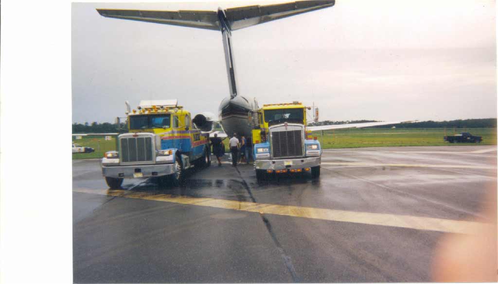 Two tow trucks pulling an airplane on a wet runway. Gray sky and grass in background.