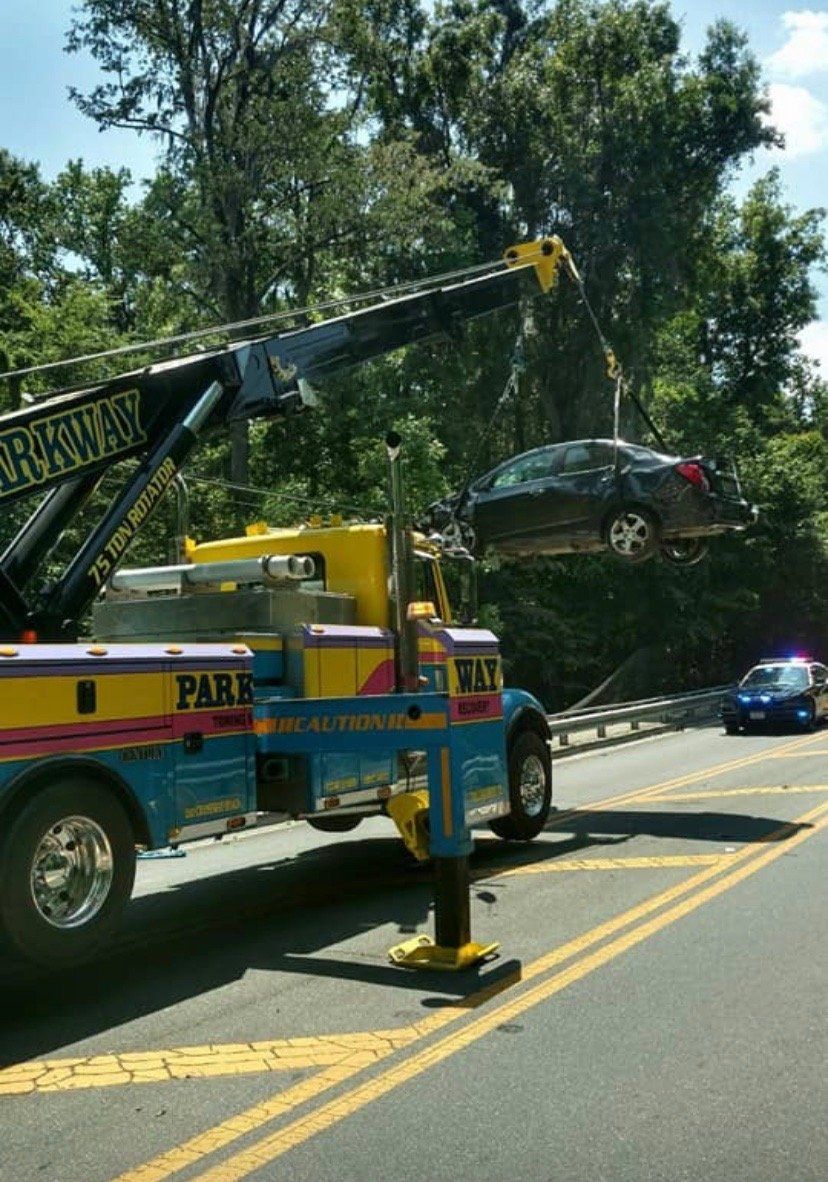 Tow truck lifting a dark sedan, parked on a road with yellow lines, in a wooded area.