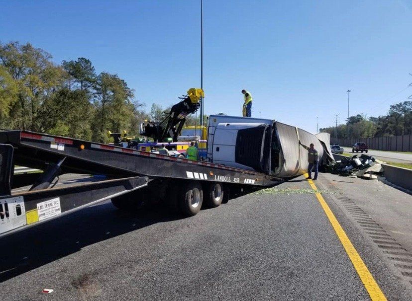 Flatbed trailer with a tipped-over cargo container on the side of a highway; two workers near the scene.