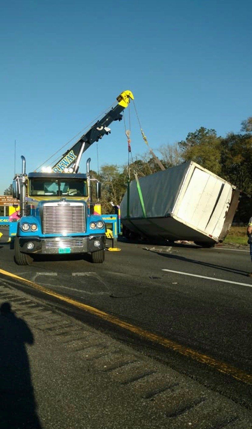 A blue tow truck lifting a tipped-over white cargo container on a highway with a blue sky.