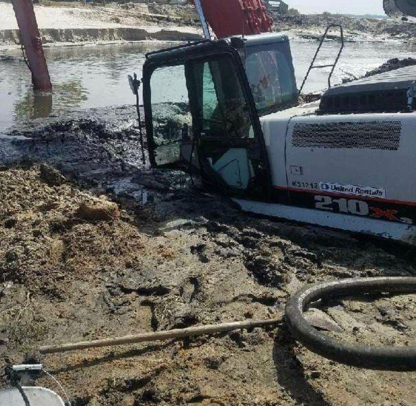Excavator stuck in mud near a body of water. Bucket, cab, and some wheels submerged.