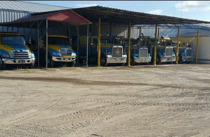 Tow trucks parked under a metal roofed shelter on a gravel lot. Trucks are blue and yellow.