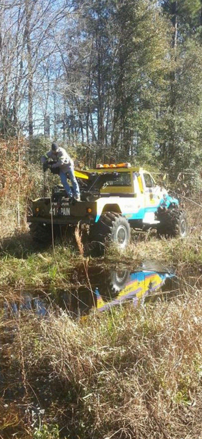 A tow truck is pulling a vehicle from a body of water in a grassy field, with a person working on the truck.
