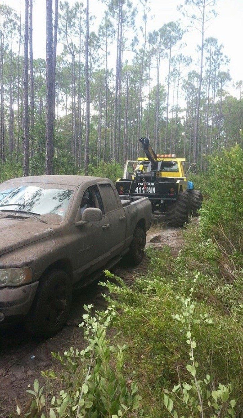 Muddy truck being towed by a yellow off-road vehicle through a wooded area.