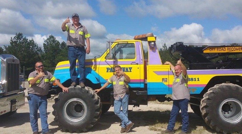 Four people pose with a brightly colored monster truck, giving thumbs up; outdoor setting, sunny day.