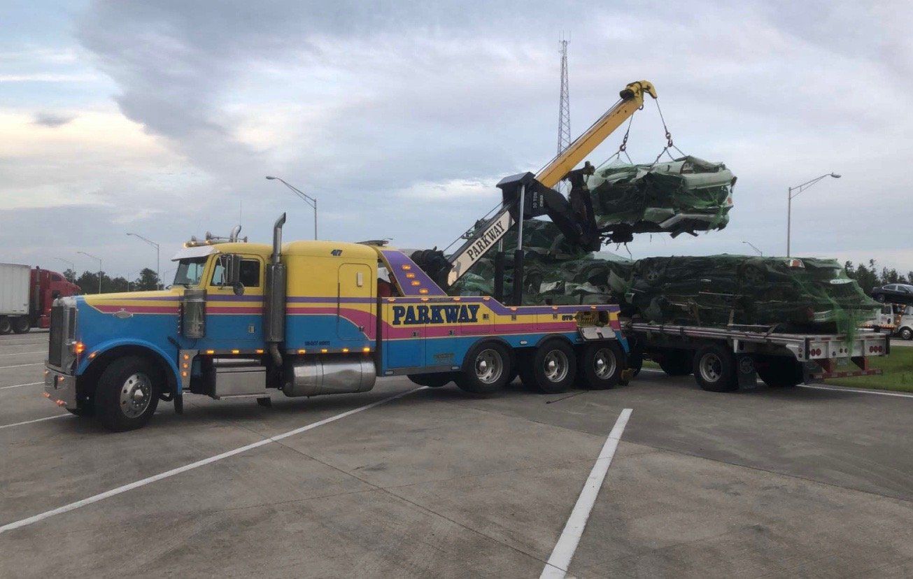 A tow truck lifts a bundle of green-wrapped items from a flatbed trailer in a parking area.