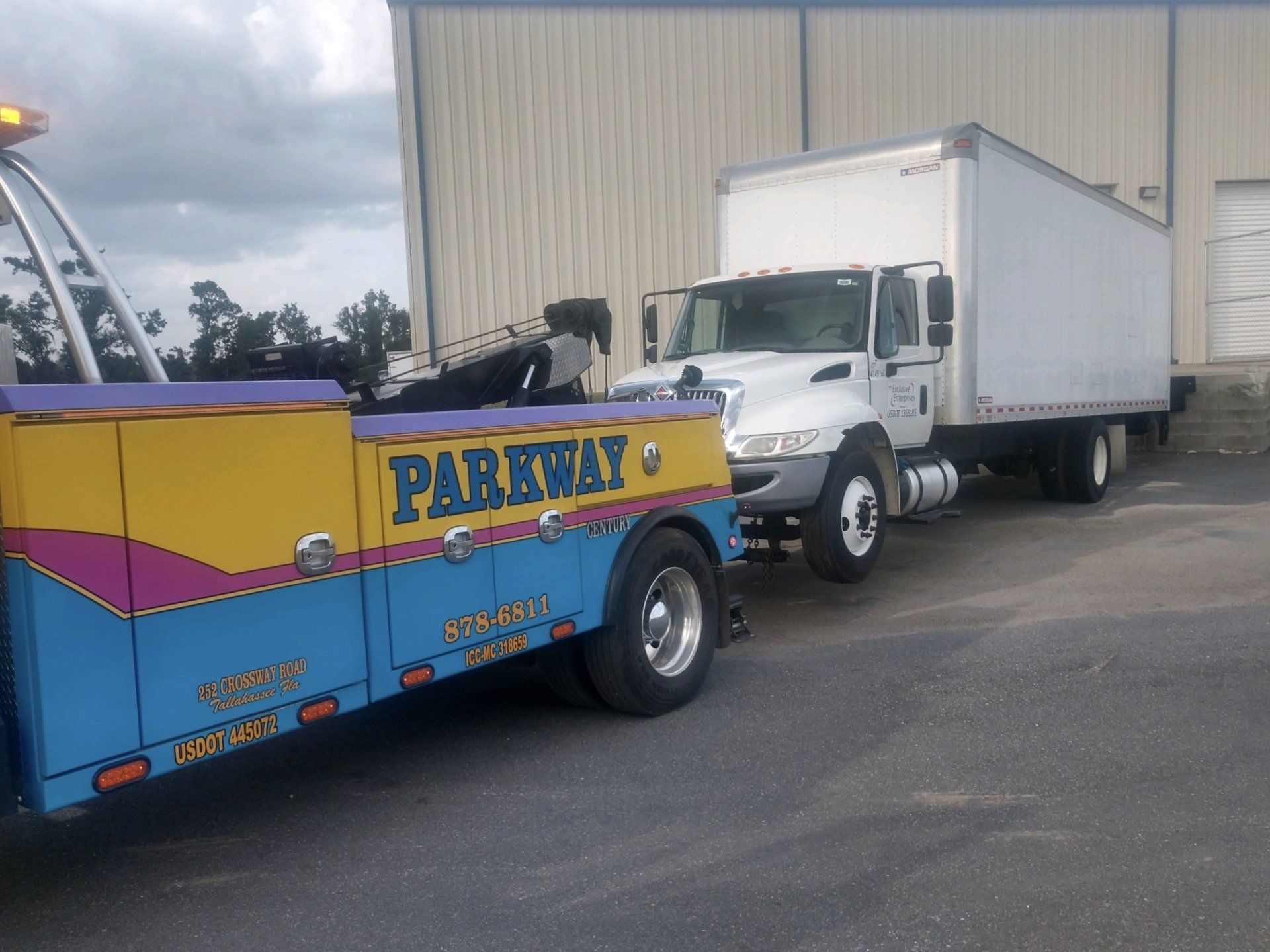 Tow truck towing a white box truck. The tow truck is blue, yellow, and purple. Building in the background.