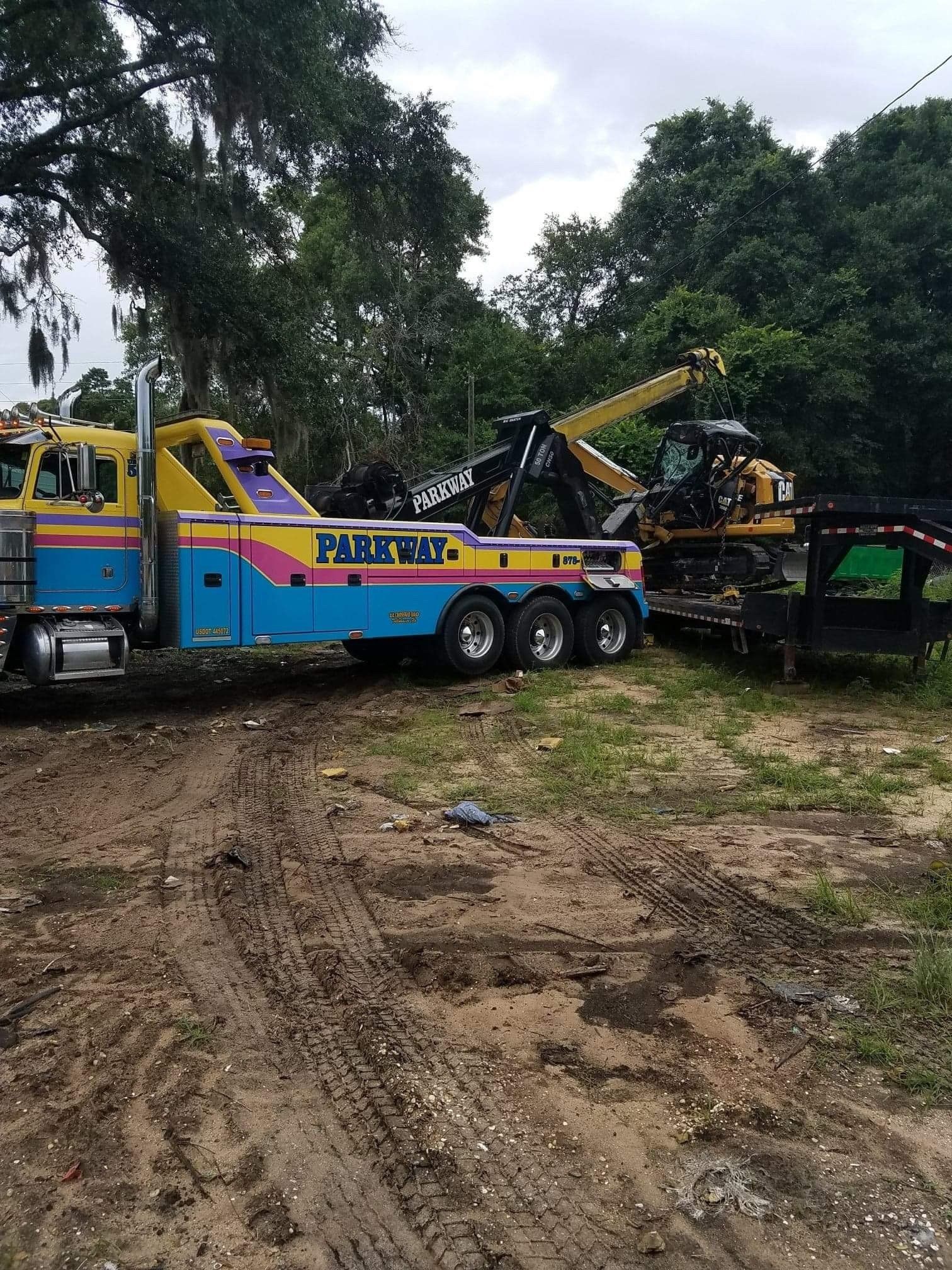 Tow truck loading heavy equipment onto a trailer on a muddy ground. Parkway logo on the truck.