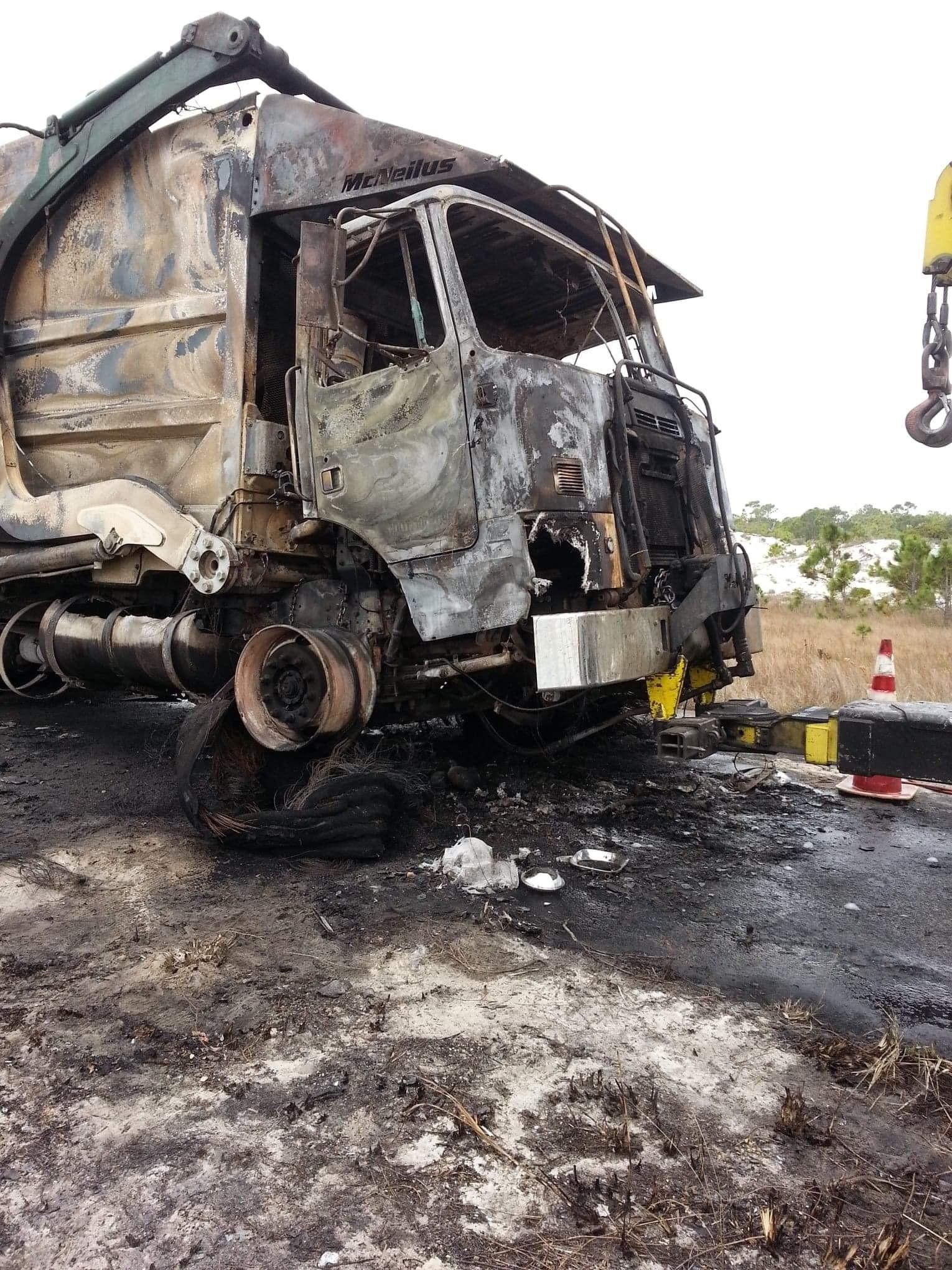Burned out truck on a road; charred exterior, missing tires, being lifted by crane.