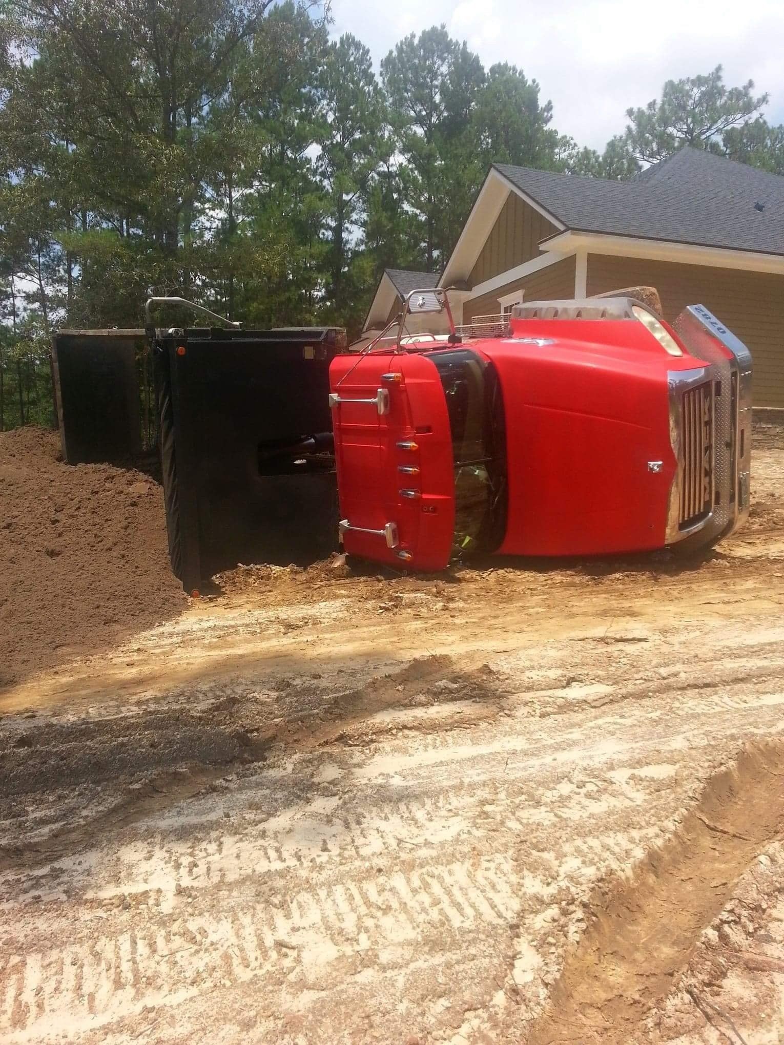 Red semi-truck overturned next to a dirt pile and a light brown building on a construction site.