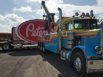 Tow truck with a large Peters sign on a flatbed trailer. Blue, yellow, and purple truck. Bright outdoor setting.