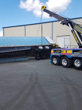 Tow truck lifting a semi-trailer that has overturned; outdoor setting with a building and blue sky.