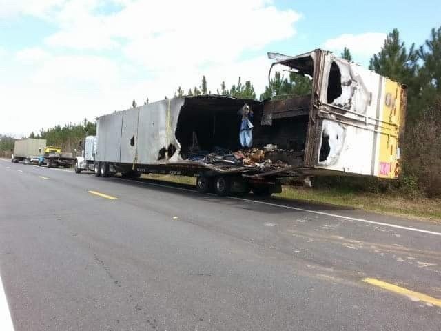Burned semi-truck trailer on side of road with visible fire damage. Other vehicles present.
