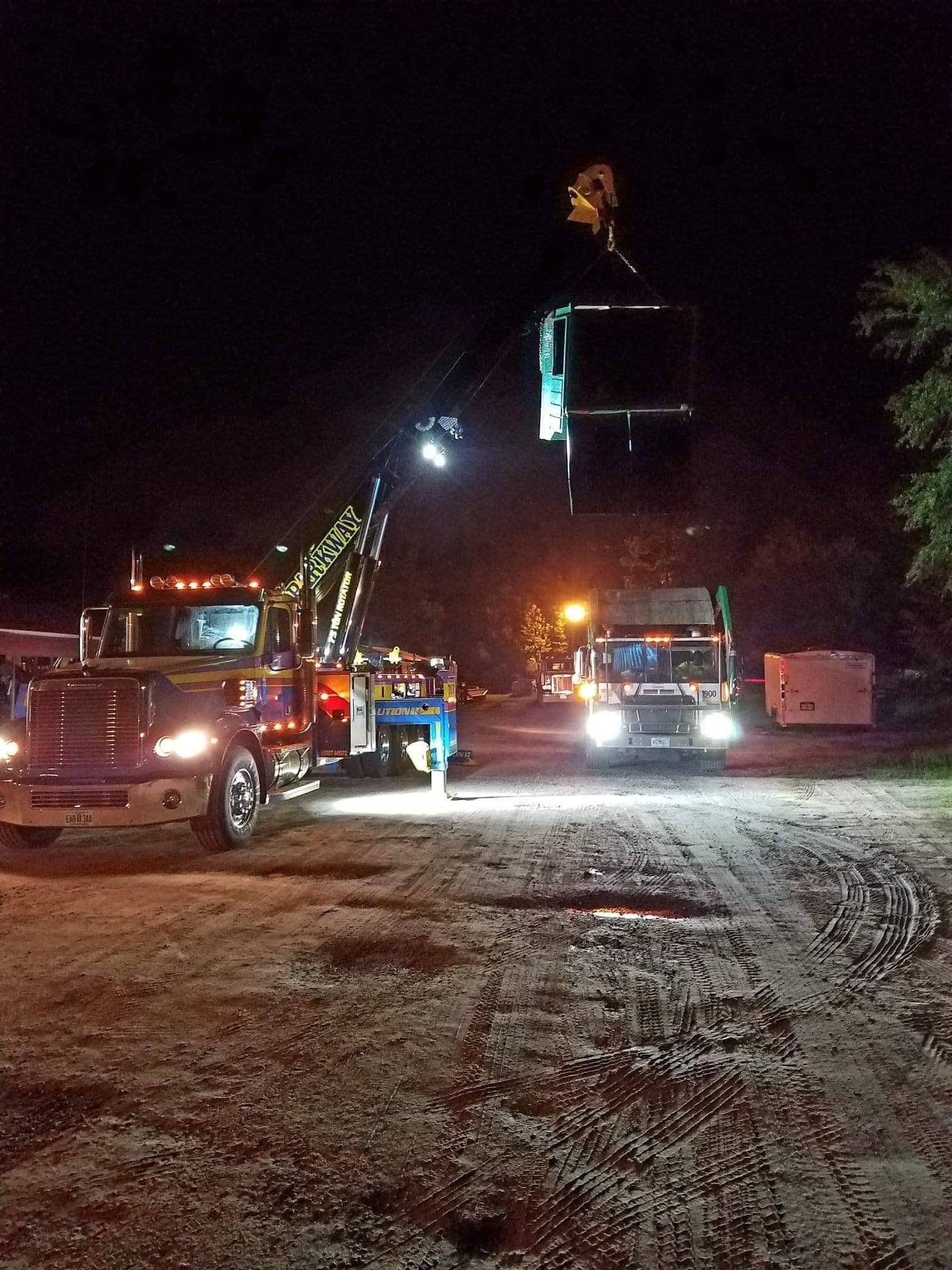 Tow truck lifts a container at night. Another truck, lights on, parked nearby on a dirt road.