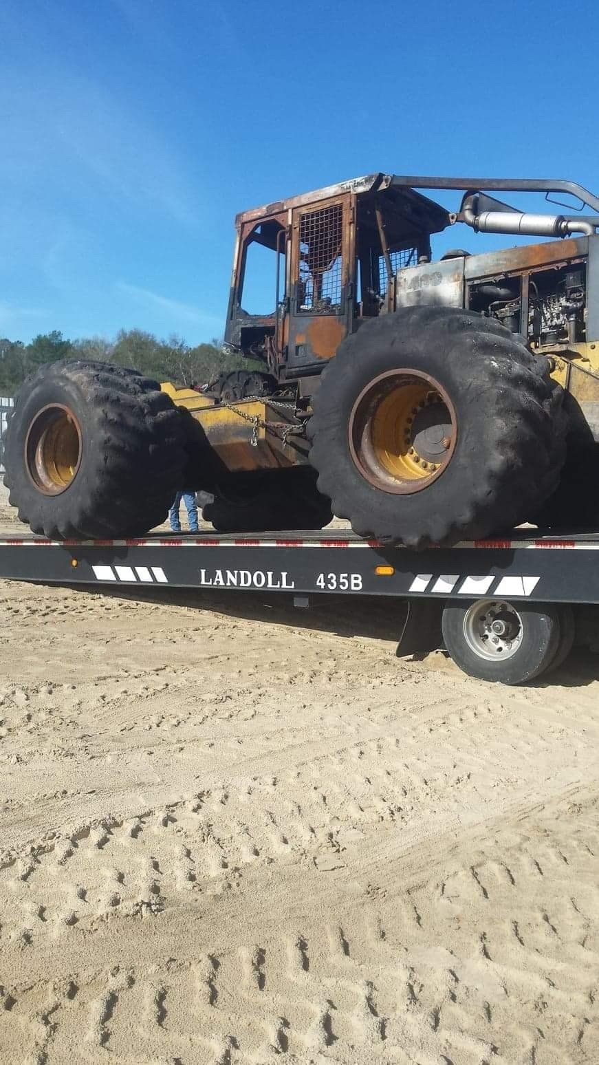 Burned yellow logging machine on a flatbed trailer, outdoors.