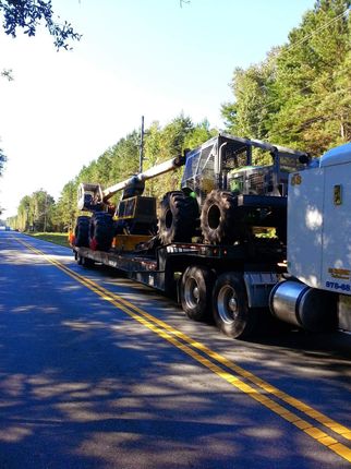 Semi-truck hauling heavy machinery on a highway; sunny day, trees in background.