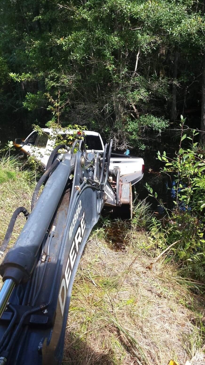 Excavator arm in foreground, retrieving a white truck from a wooded ditch.