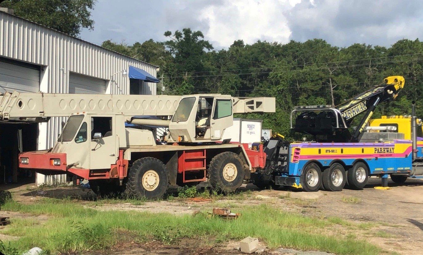 A large crane being towed by a blue and yellow tow truck outside a building with trees in the background.