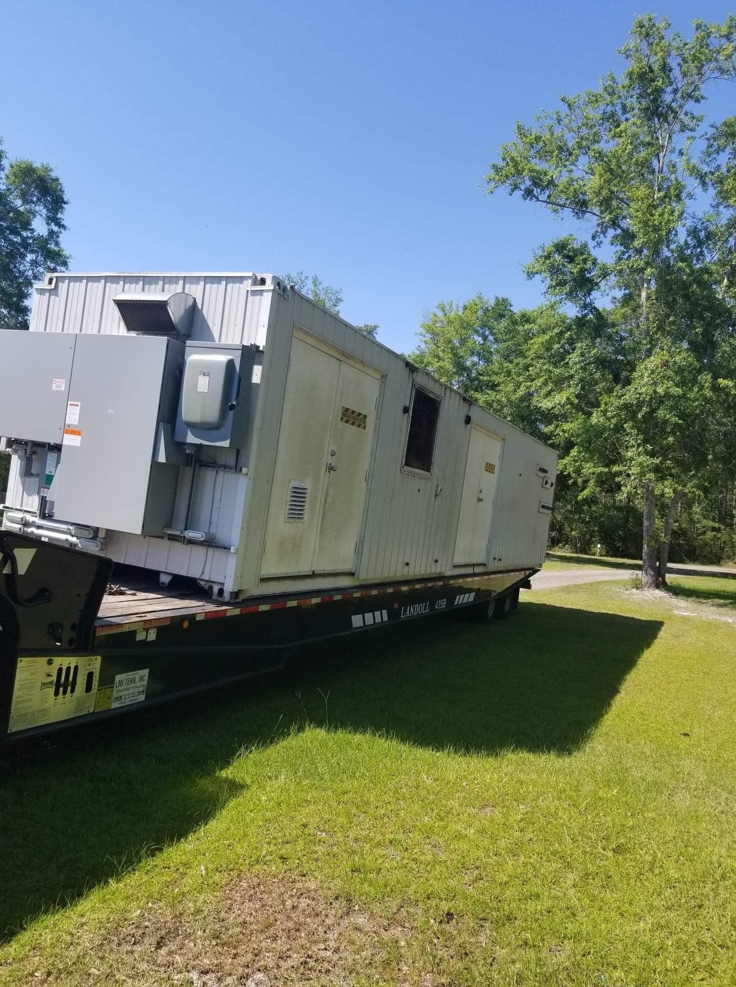 A large, industrial generator unit on a flatbed trailer, parked on grass under a clear, sunny sky.