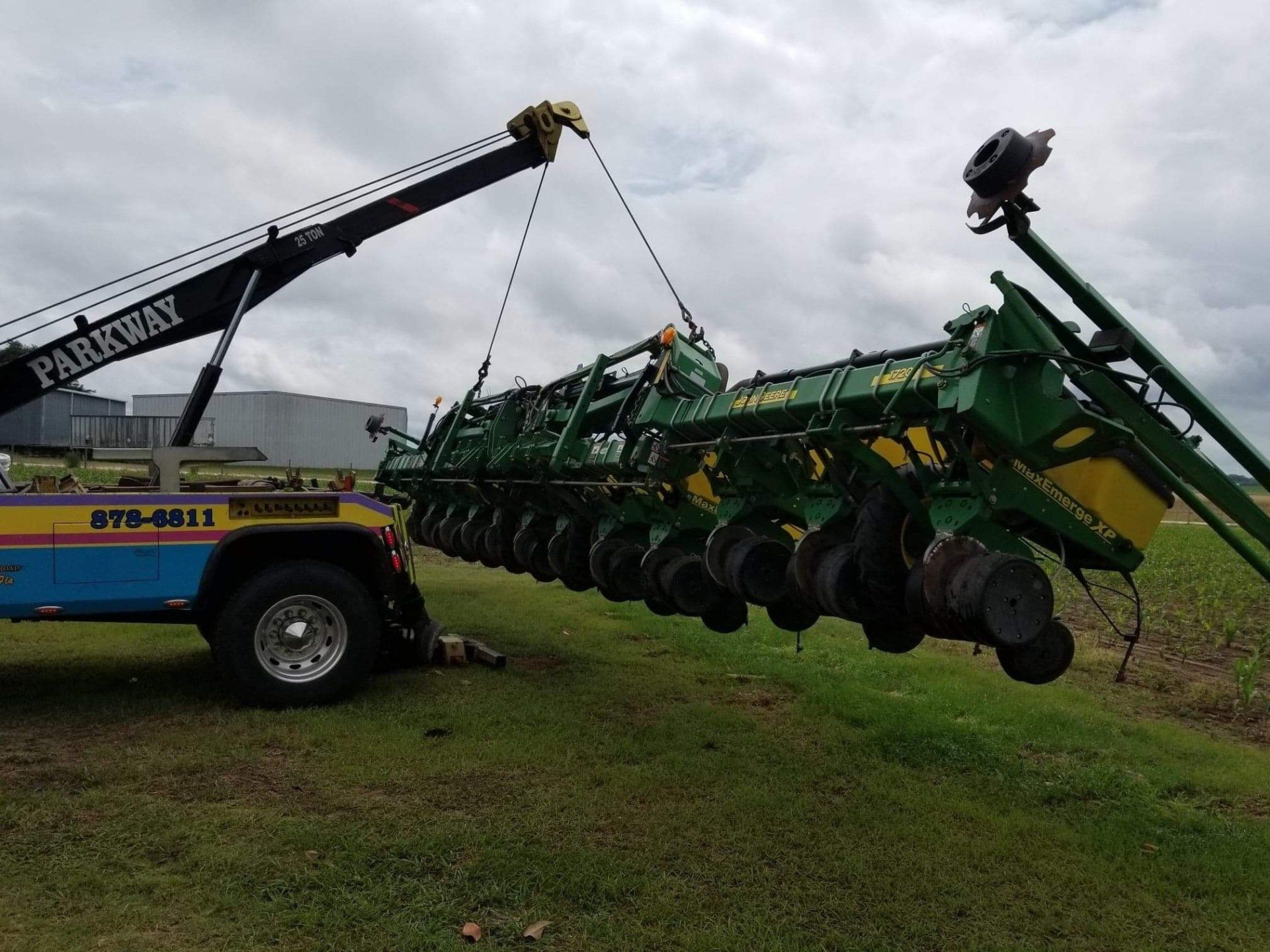 Tow truck lifting a large green John Deere planter in a field on a cloudy day.