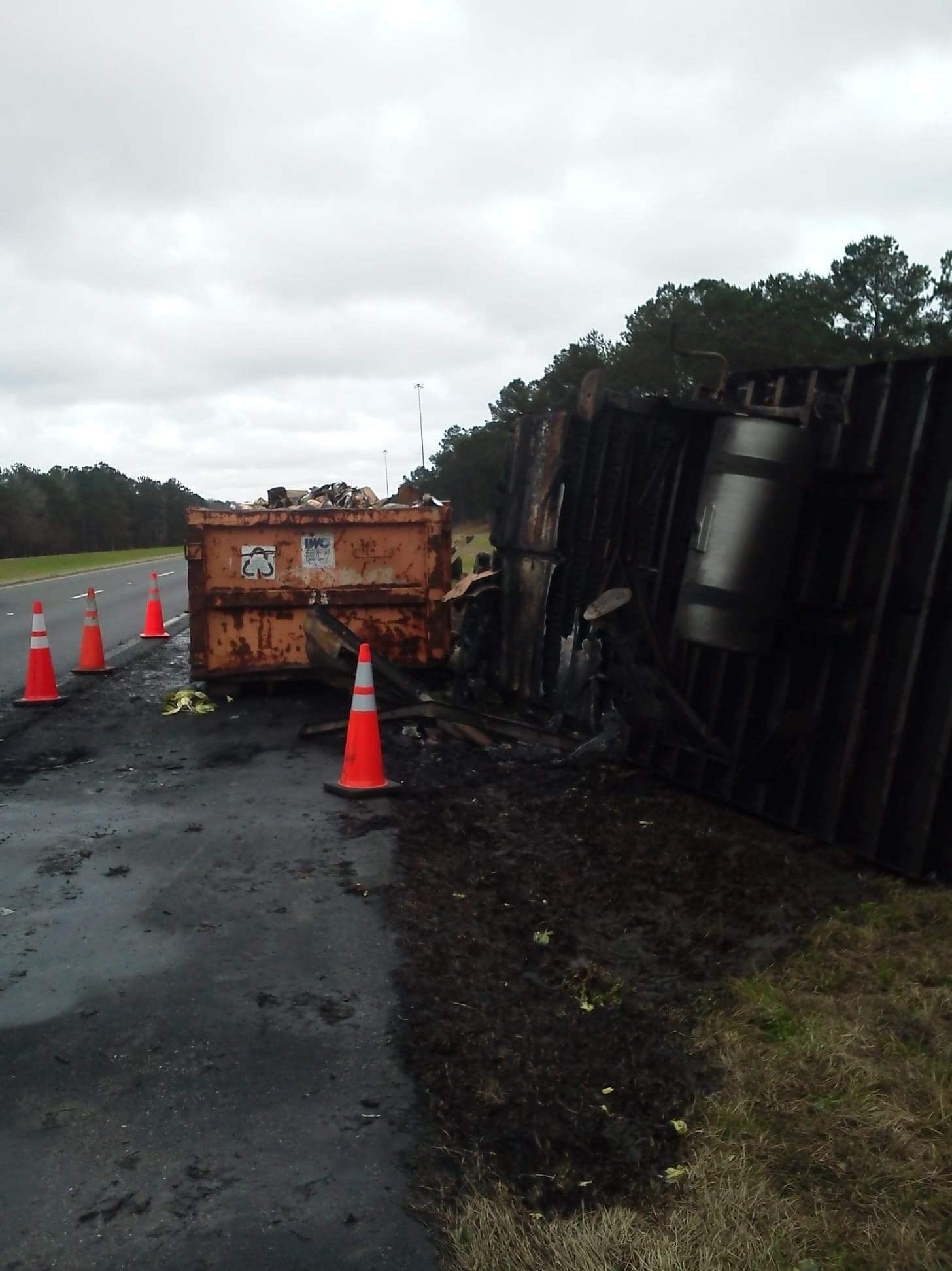 A damaged semi-truck trailer on its side next to a highway. Debris and traffic cones surround it.