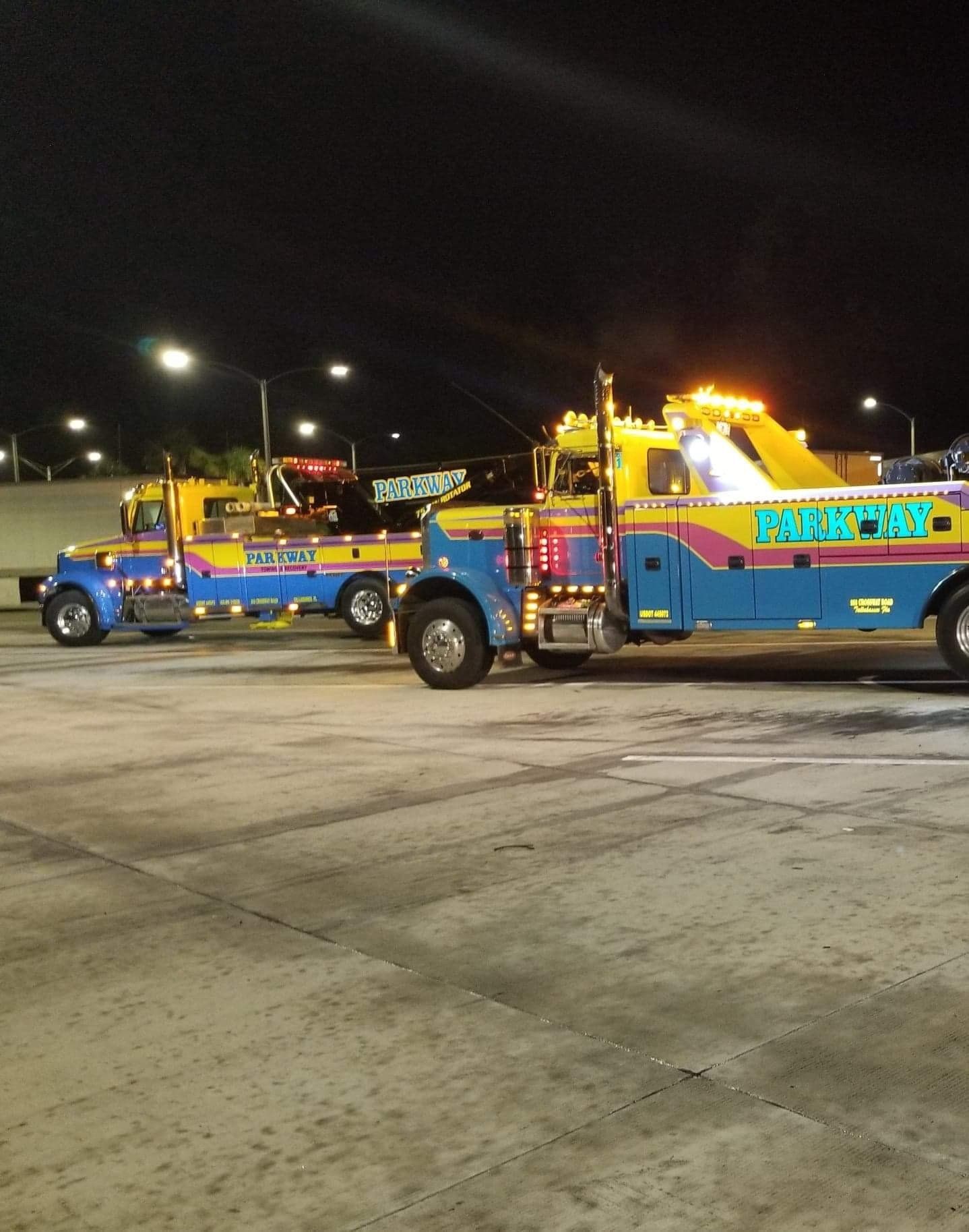 Two colorful tow trucks with flashing lights parked at night.