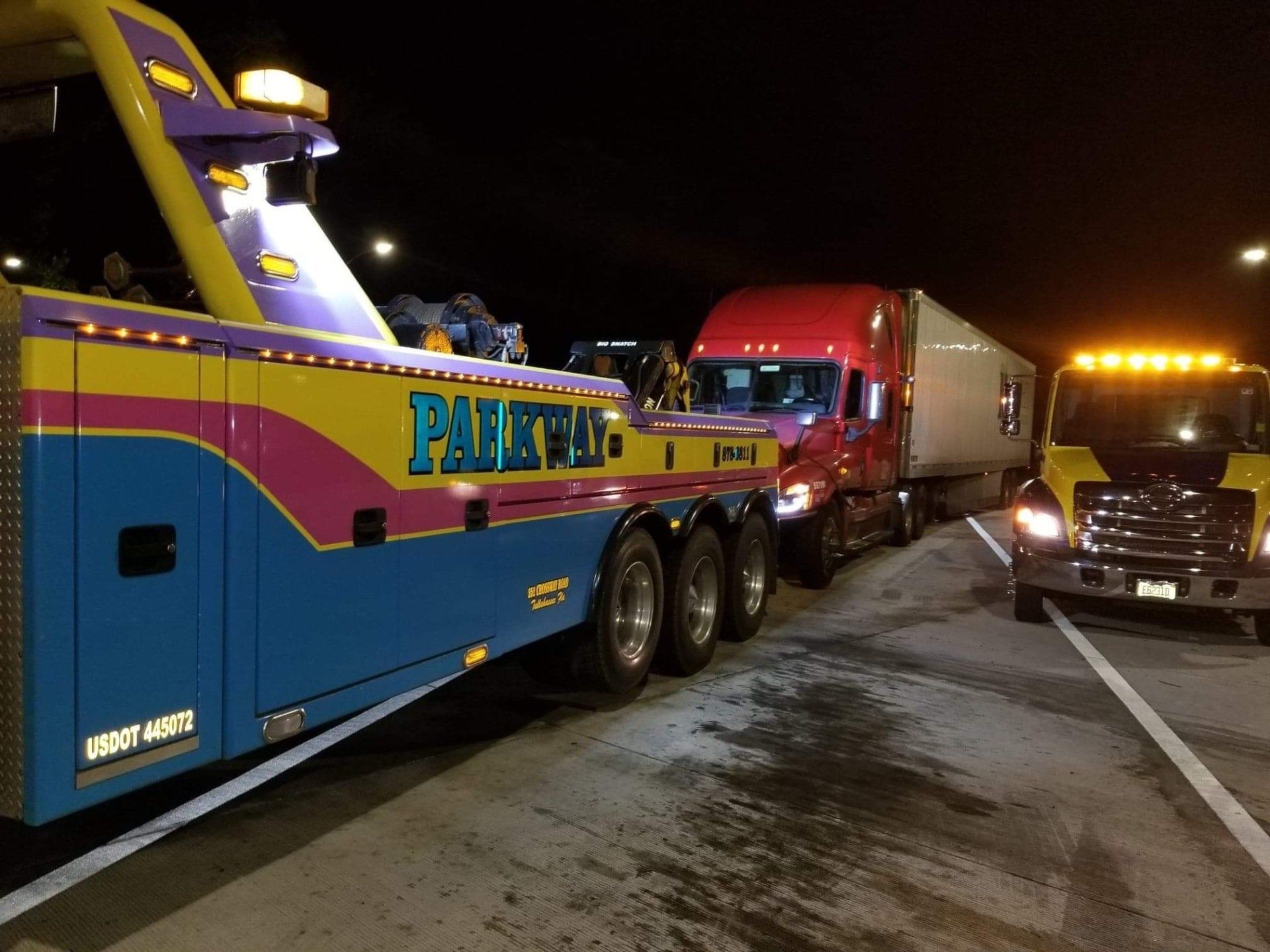Tow trucks preparing to tow a semi-truck at night. One tow truck is colorful, another is yellow with lights on.