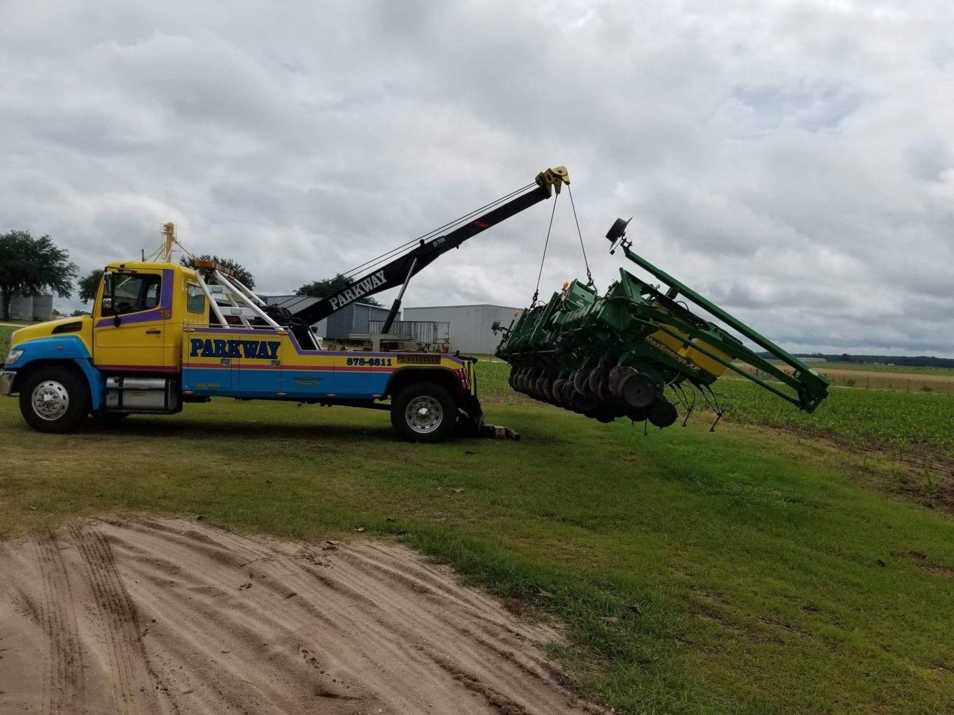 Tow truck lifting a green agricultural machine in a field. The truck is yellow and blue, parked on grass.