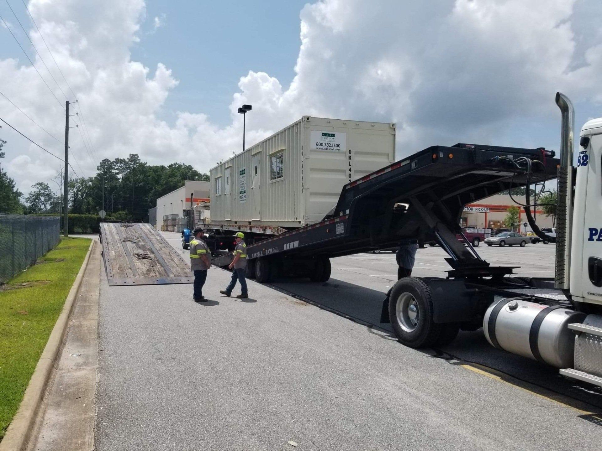 A semi-truck unloading a cargo container at a paved area with two workers assisting.