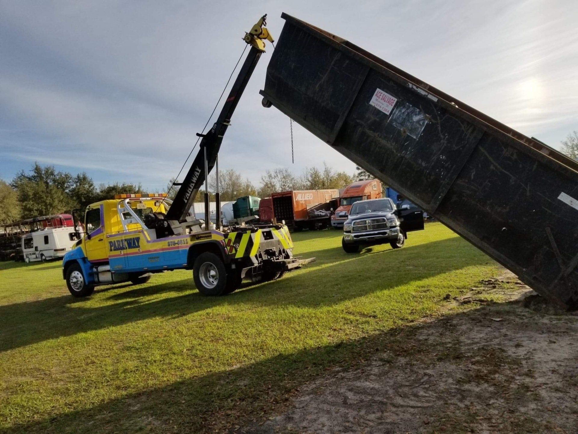 Tow truck lifting a dumpster in a grassy area with vehicles in the background.
