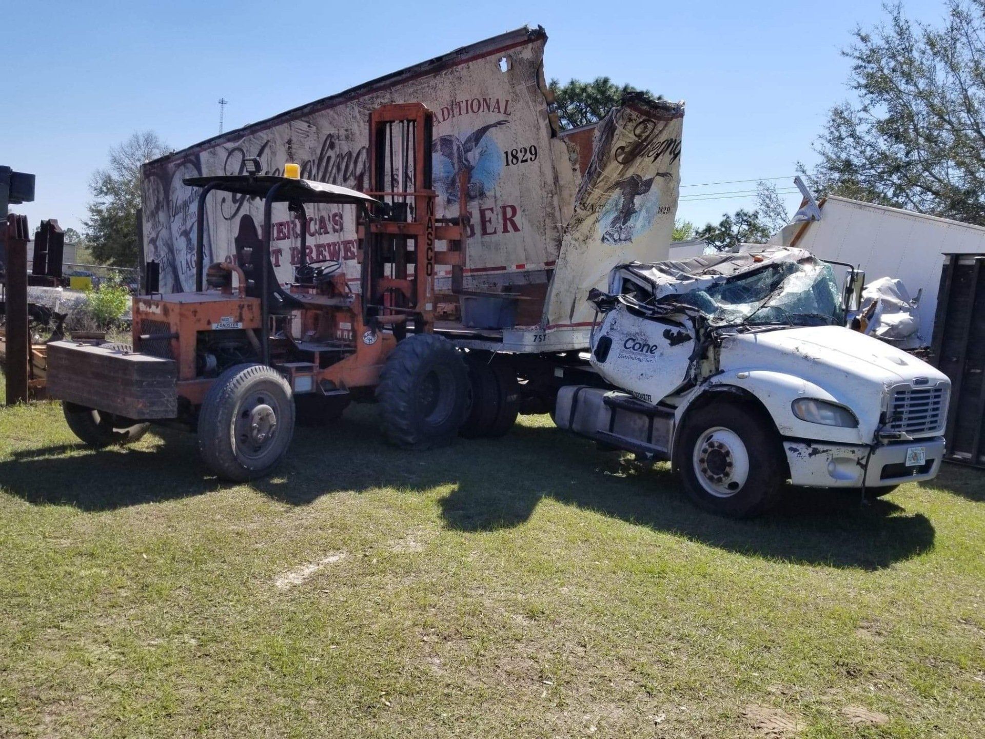 An orange forklift and a damaged white truck are next to a weathered building with signage.