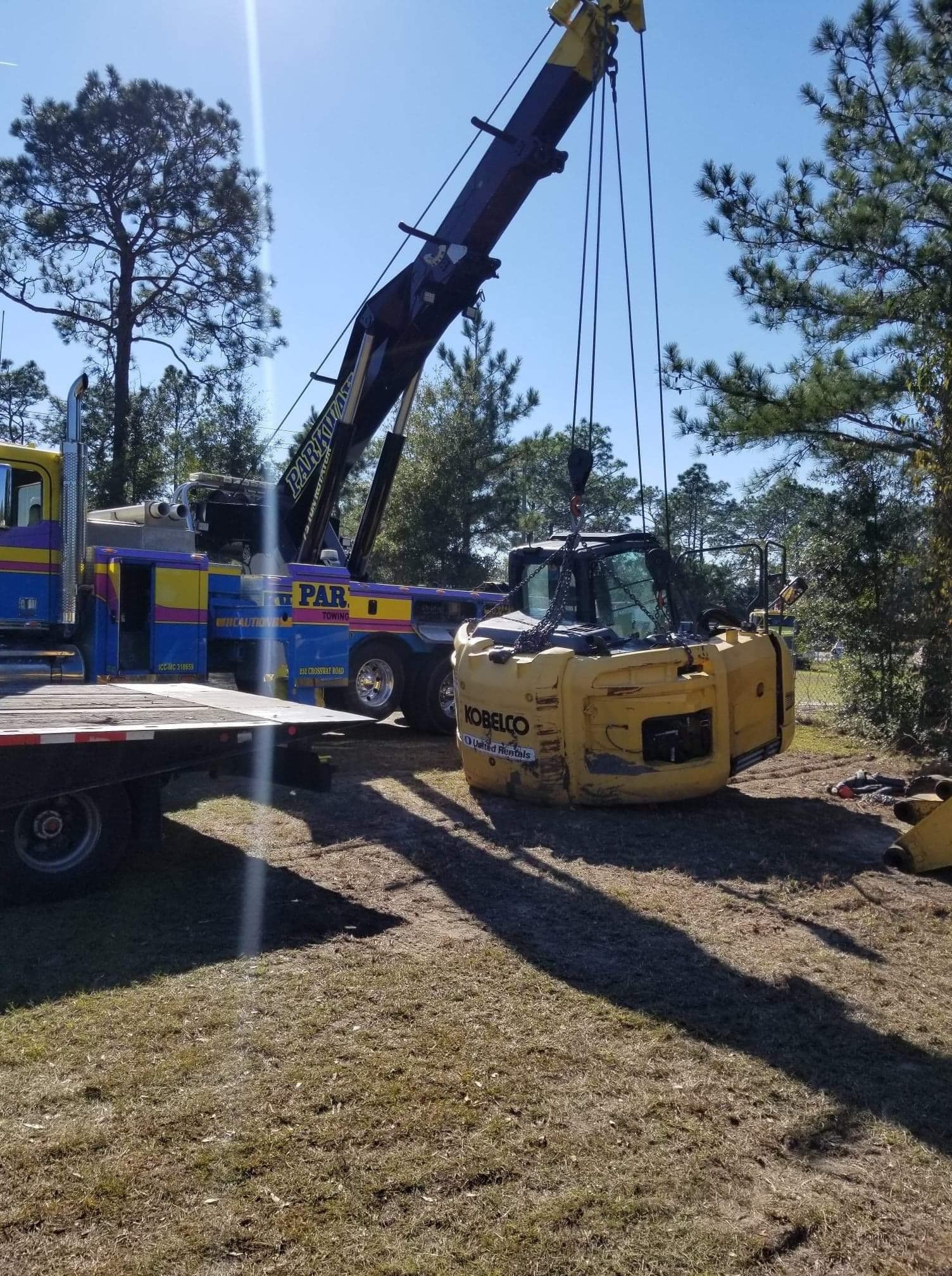 A yellow excavator being lifted by a crane onto a flatbed trailer in a grassy area.