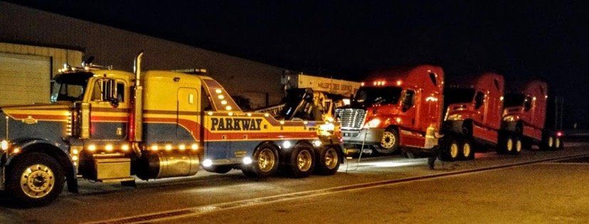 A tow truck is towing multiple red semi-trucks at night. The tow truck is yellow and blue, and has 