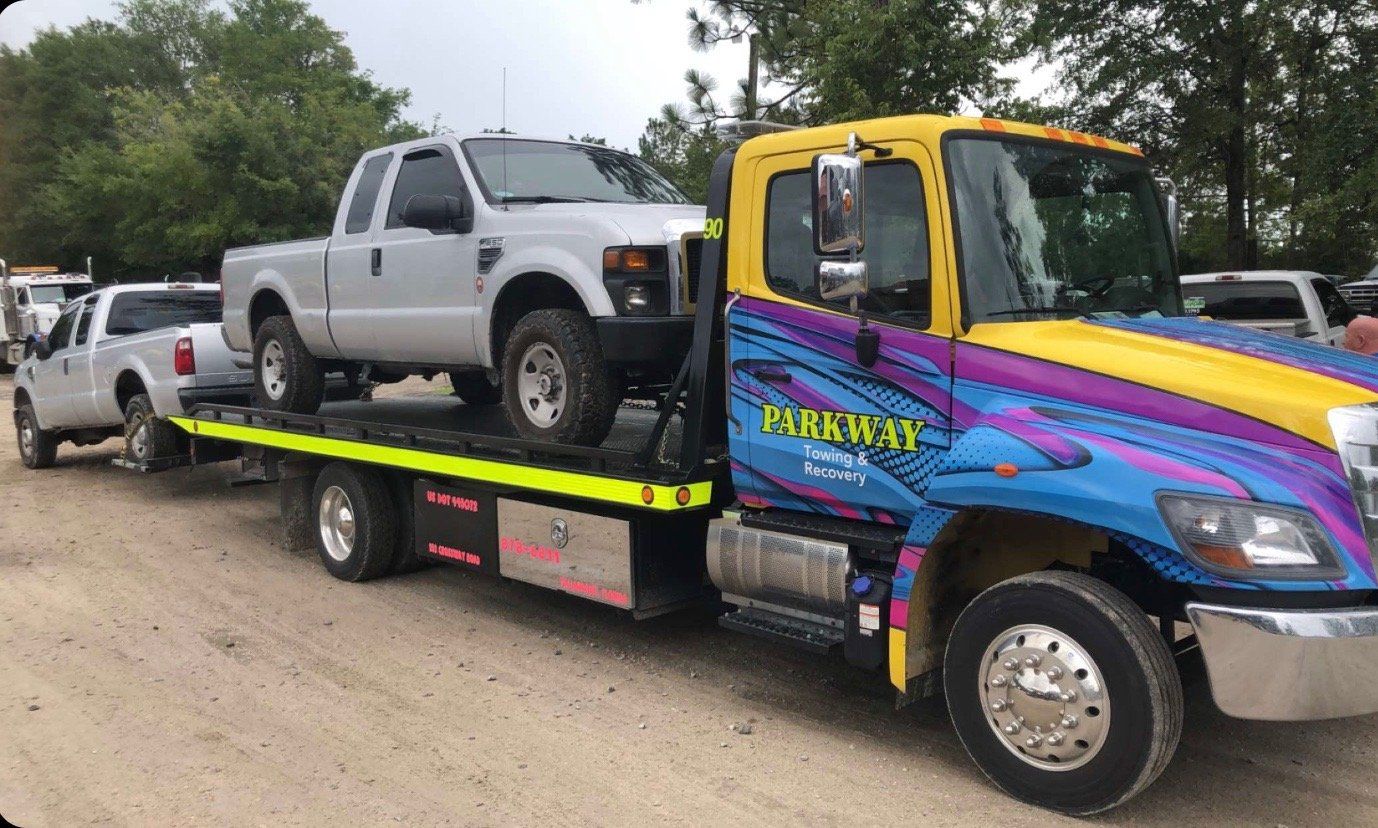 Tow truck with a silver pickup truck on its flatbed and another being towed on a dirt road.