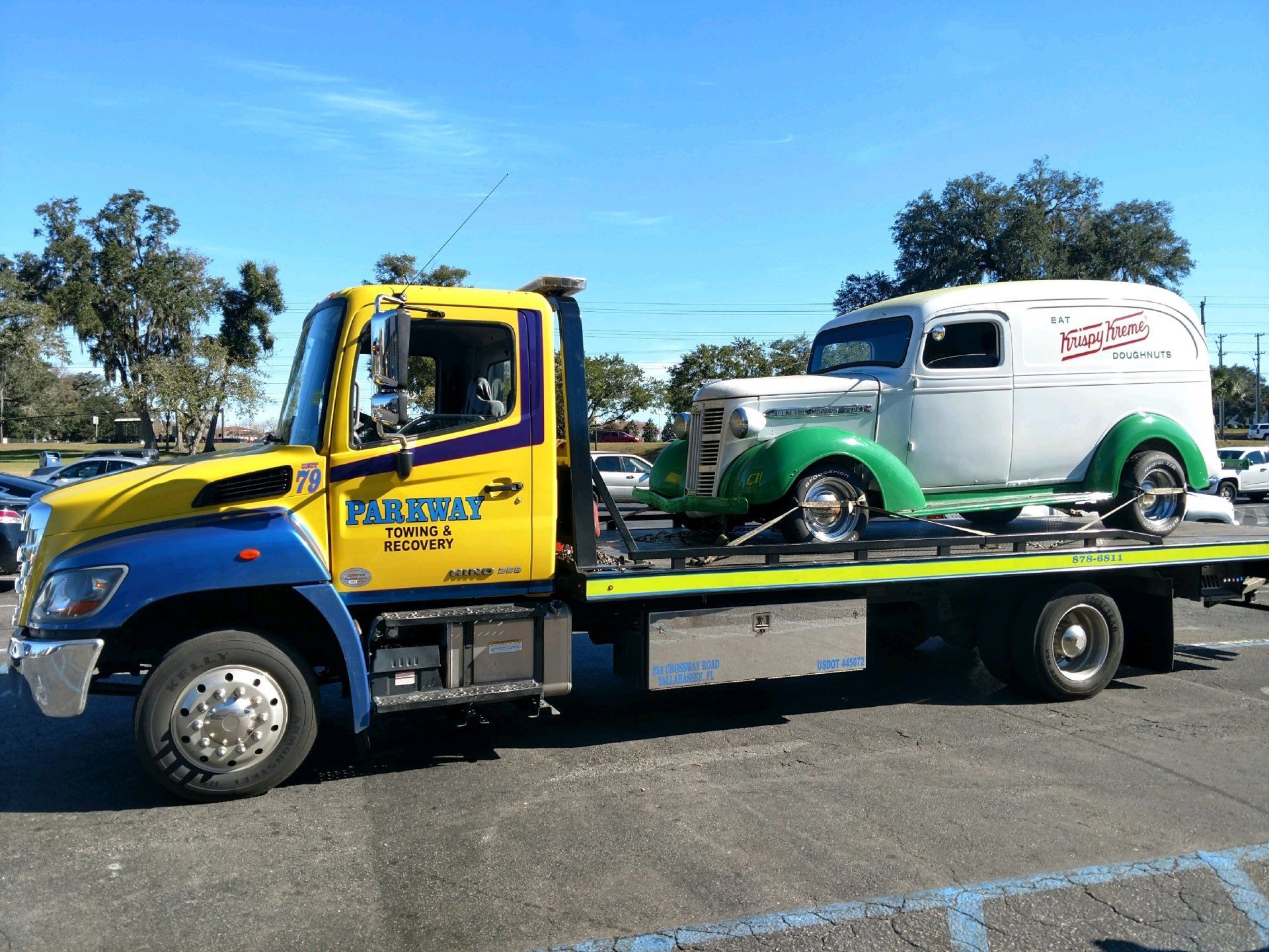 Tow truck with a white and green vintage truck on its flatbed. Sunny day, outdoor setting.