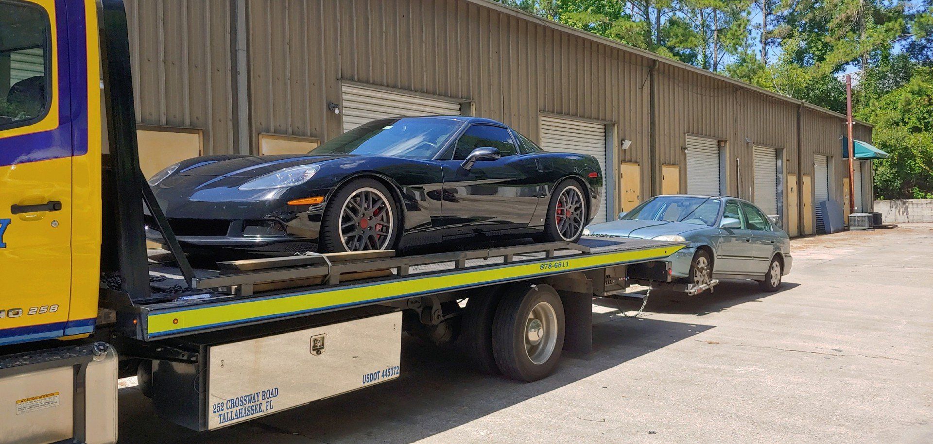 Black Corvette loaded on a tow truck, towing a gray sedan in front of a warehouse.