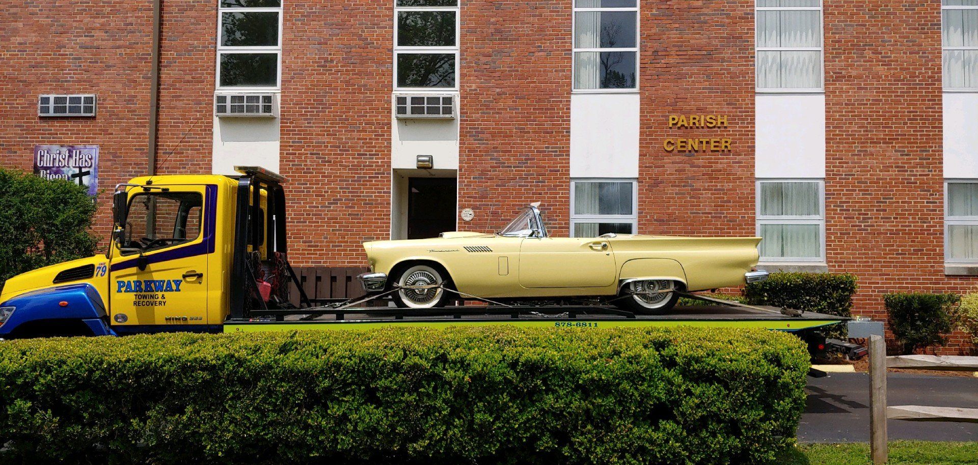 A yellow tow truck carries a light yellow vintage convertible car, parked in front of a brick building.