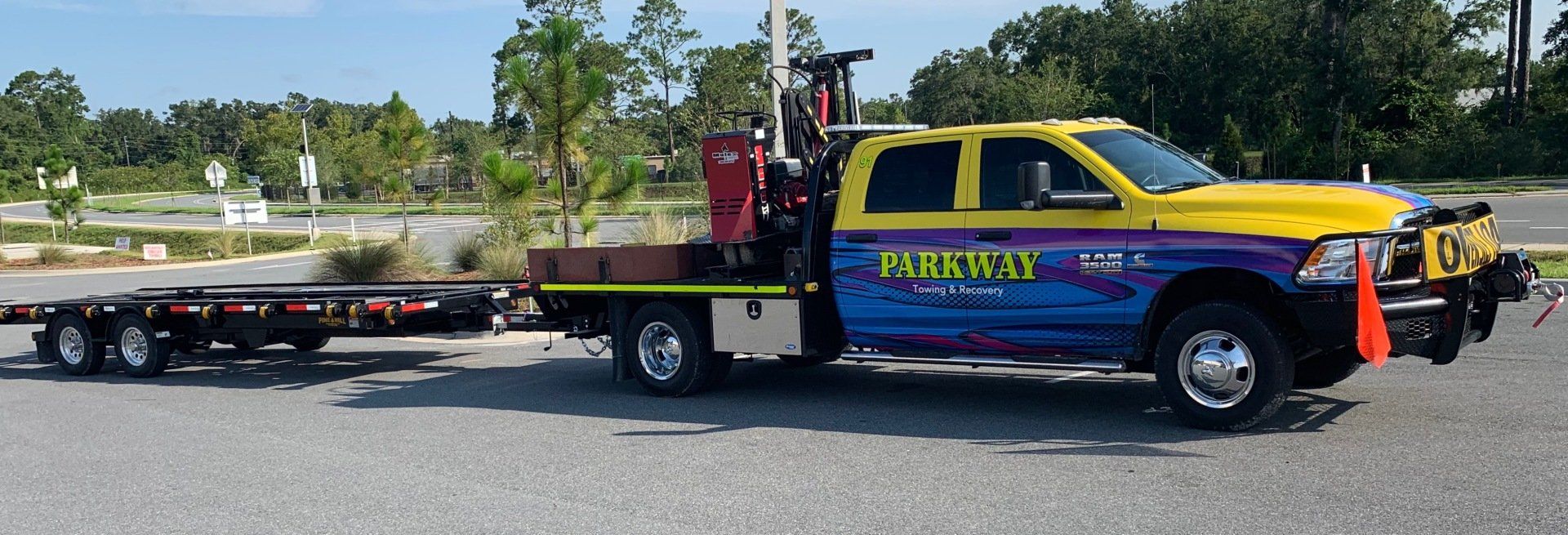 Yellow and blue Parkway tow truck with an attached trailer, parked on pavement. Trees and sky in the background.
