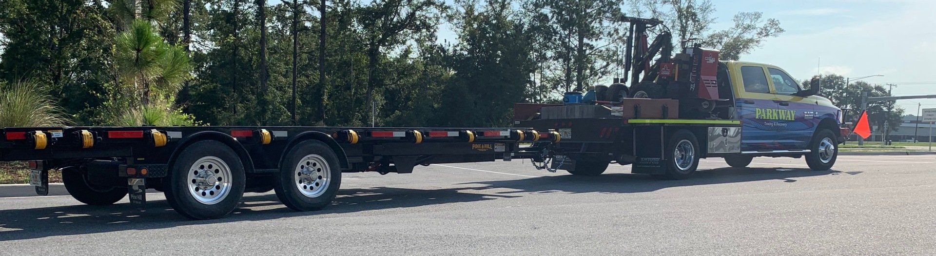 Truck towing a flatbed trailer on a gravel road, with trees in the background under a blue sky.