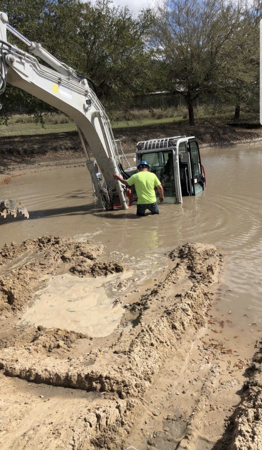 A man stands in muddy water next to a submerged construction vehicle; excavator arm reaches over.