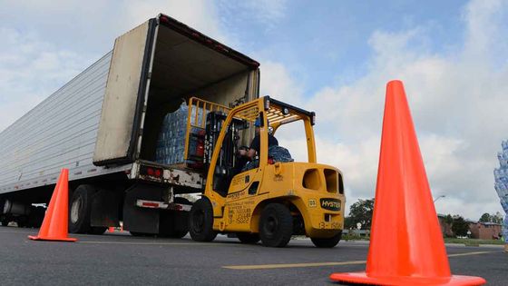 Yellow forklift loading bottled water into a semi-truck. Orange cones on asphalt. Blue sky in the background.