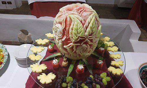 Buffet con un vassoio di vetro ,una anguria al centro con disegni di fiori e intorno uva nera,uva bianca e fette di ananas
