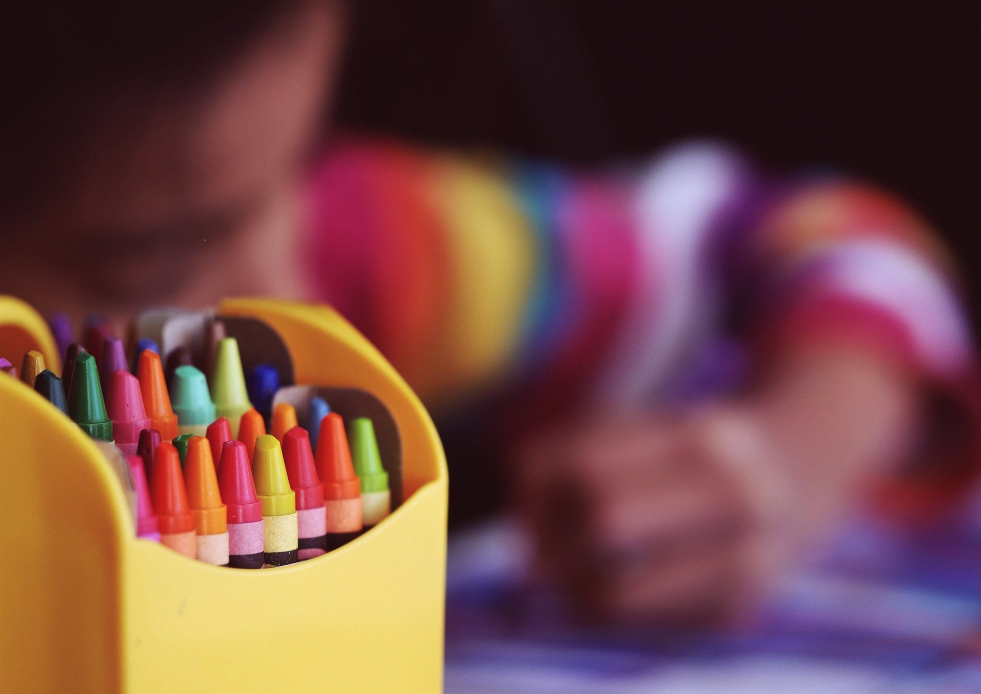 Yellow crayon box with various colored crayons, child in striped shirt drawing in background.