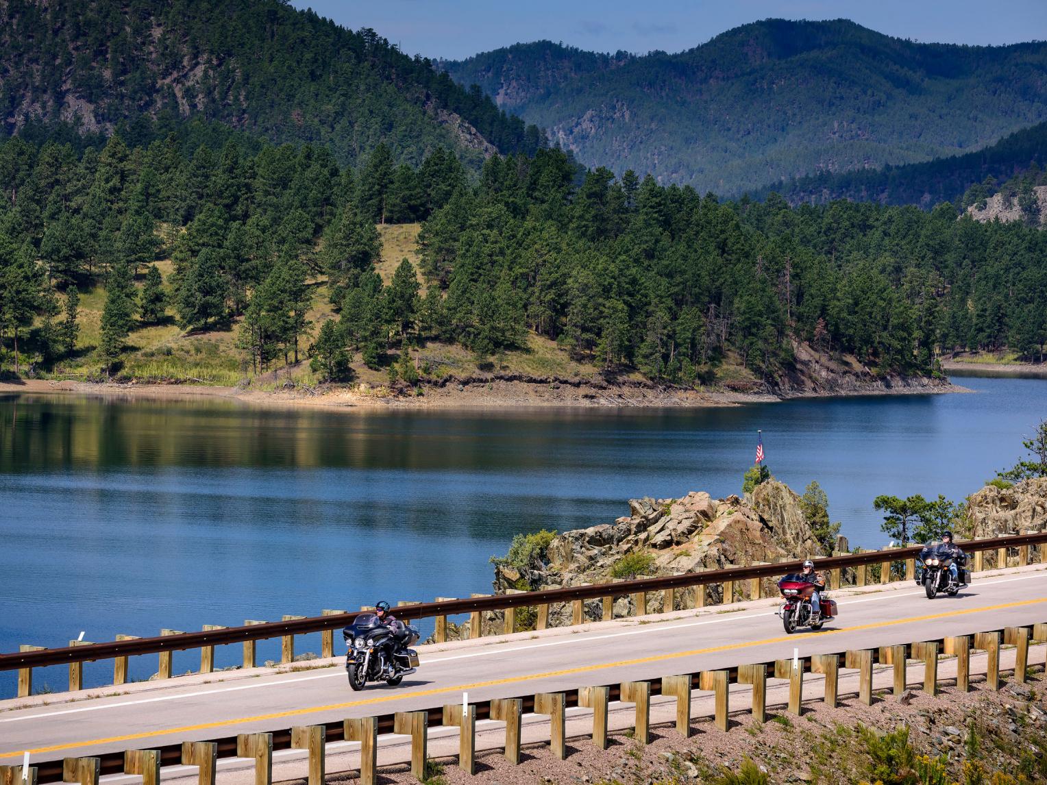 Two people are riding motorcycles on a bridge over a lake.
