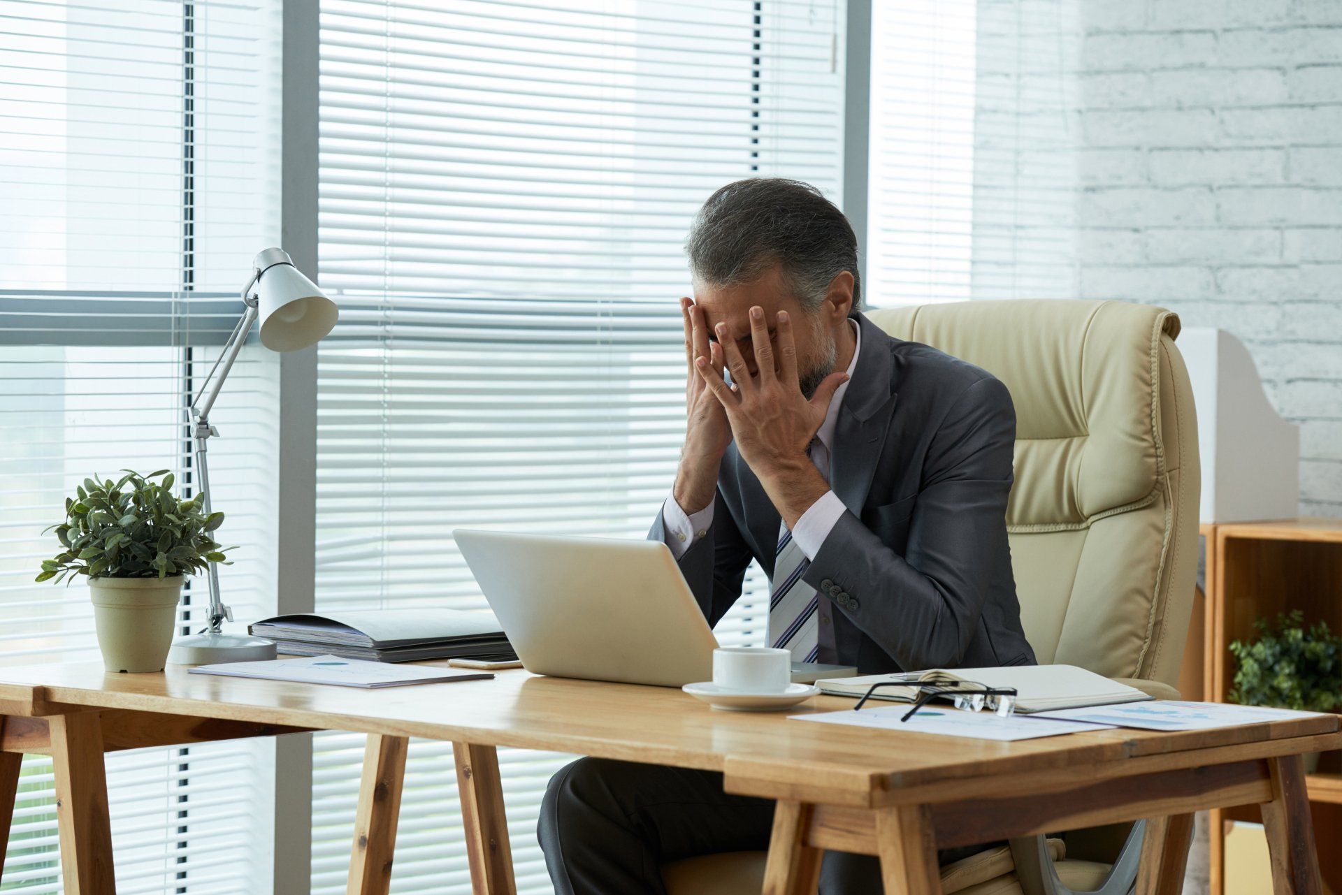 Hombre de traje estresado, cubriéndose la cara en un escritorio con una computadora portátil, entorno de oficina.