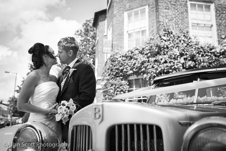 A black and white photo of a bride and groom kissing in front of a wedding car